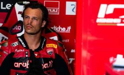 Male motorcyclist in a red Ducati racing suit sits in a sponsor-filled pit area, looking off to the side.