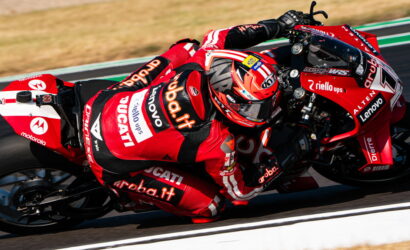 Red Ducati racing motorcycle with rider leaning into a curve, sponsor logos visible (aruba.it, Lenovo, Motorola) on a racetrack.