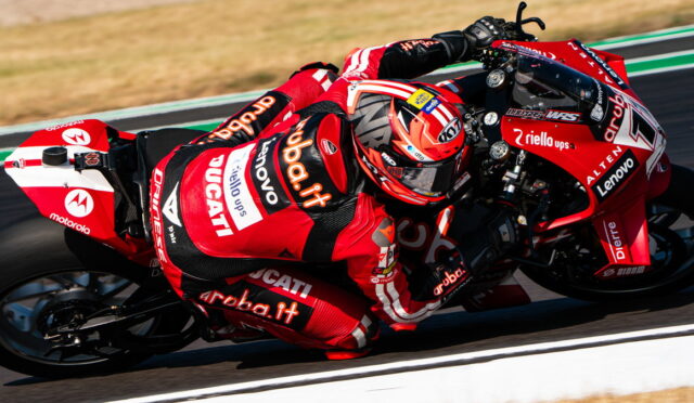 Red Ducati racing motorcycle with rider leaning into a curve, sponsor logos visible (aruba.it, Lenovo, Motorola) on a racetrack.