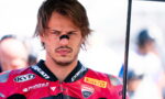 Male motorcycle racer in a red Ducati racing suit with sponsor logos, looking focused, holding an umbrella.