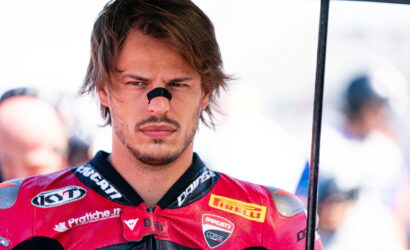 Male motorcycle racer in a red Ducati racing suit with sponsor logos, looking focused, holding an umbrella.