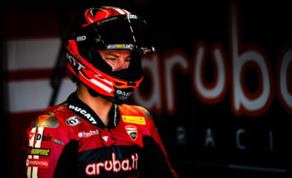 Moto racing rider in red Ducati suit and helmet, looking to the left in a dim pit area with a blurred backdrop showing 'Aruba.it'
