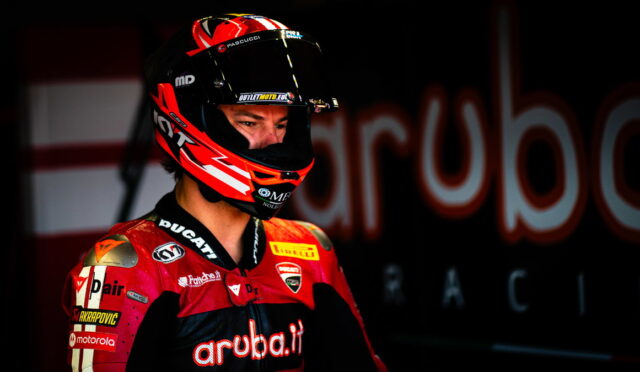 Moto racing rider in red Ducati suit and helmet, looking to the left in a dim pit area with a blurred backdrop showing 'Aruba.it'