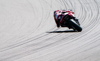 Motorcyclist in a red racing suit leans into a sharp left turn on a race track, tire marks visible on the pavement.