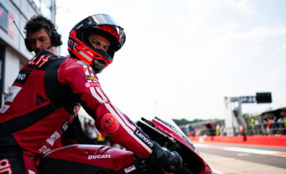 Professional motorcycle racer in a red Ducati racing suit sits on a red Ducati motorcycle in the pit lane before a race, wearing a helmet with visor up slightly.