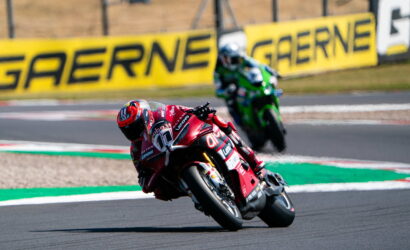 Red racing motorcycle leaning hard into a corner on a racetrack, a green bike behind, with bright GAERNE banners in the background.