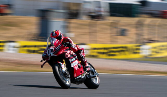 Motorcycle racer in a red racing suit leans into a curve on a race track with motion blur in the background.