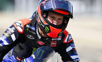 Professional motorcycle racer wearing a full racing suit and helmet, leaning into a turn with sponsor logos visible on the suit and bike.