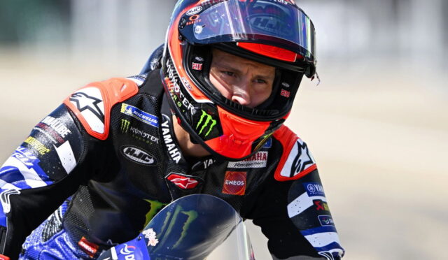 Professional motorcycle racer wearing a full racing suit and helmet, leaning into a turn with sponsor logos visible on the suit and bike.