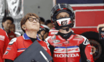 Staff member with a clipboard beside a rider in a red Honda racing suit and helmet, both looking upward in a pit area.