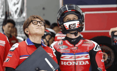 Moto racer in red Honda suit wearing helmet stands beside a team member holding a clipboard, both looking upward in a pit area.