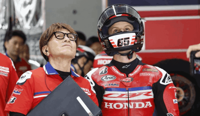 Moto racer in red Honda suit wearing helmet stands beside a team member holding a clipboard, both looking upward in a pit area.