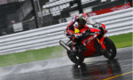 Motorcycle racer in red gear leaning into a turn on a red racing bike on a wet track with spray behind the tires.