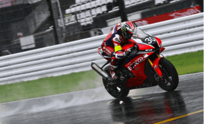 Motorcycle racer in red gear leaning into a turn on a red racing bike on a wet track with spray behind the tires.