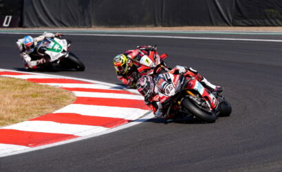 Motorcycle racers lean into a sharp turn on a race track, red-and-white curbing in the foreground.