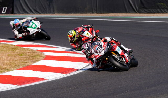 Three motorcycle racers lean into a sharp turn on a race track, riding high-speed on red-and-white curbs.