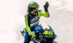 Racer on a Yamaha motorcycle, number 46, in neon yellow and blue gear with a colorful helmet, raising a hand as smoke surrounds the bike.