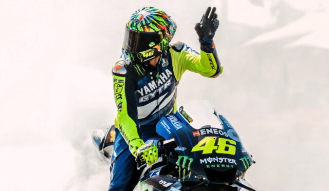 Racer on a Yamaha motorcycle, number 46, in neon yellow and blue gear with a colorful helmet, raising a hand as smoke surrounds the bike.