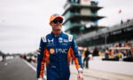 Race car driver in a blue and orange PNC racing suit and orange cap walks along the pit lane wearing sunglasses.