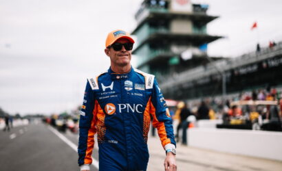 Race car driver in a blue and orange PNC racing suit and orange cap walks along the pit lane wearing sunglasses.