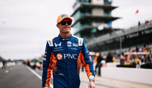 Race car driver in a blue and orange PNC racing suit and orange cap walks along the pit lane wearing sunglasses.