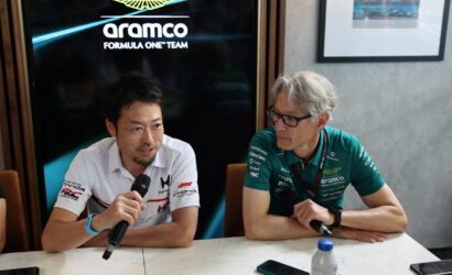 Two men sitting at a table during a press conference, one speaks into a microphone while the other listens.