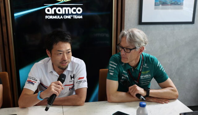 Two men sitting at a table during a press conference, one speaks into a microphone while the other listens.