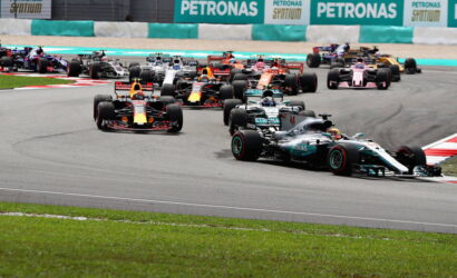 Multiple Formula 1 cars racing on a track, turning, with Petronas banners in the background. (Informative)
