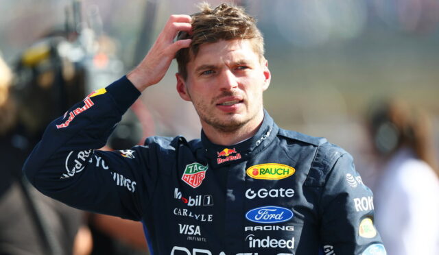 Racing driver in a dark blue Oracle suit runs his hand through his hair, standing outdoors with sponsor logos visible on his suit
