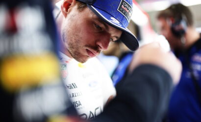 Close-up of a race driver in a blue cap, looking down as a team member adjusts his suit. Cap shows AlphaTauri branding in focus.