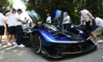 Blue race car with upward-opening doors on display, surrounded by people wearing headphones at a car show outside.