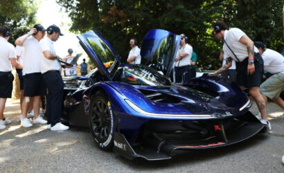 Blue race car with upward-opening doors on display, surrounded by people wearing headphones at a car show outside.
