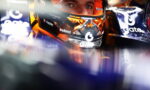 Close-up of a race car driver wearing a helmet inside the cockpit, with sponsor logos visible on the visor and car sides