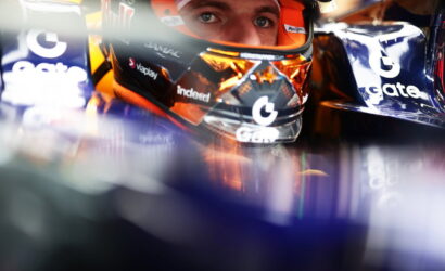 Close-up of a race car driver wearing a helmet inside the cockpit, with sponsor logos visible on the visor and car sides