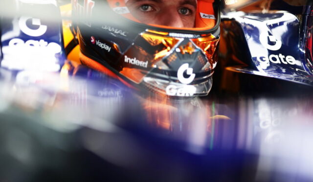 Close-up of a race car driver wearing a helmet inside the cockpit, with sponsor logos visible on the visor and car sides
