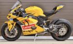 Yellow MotoGP racing motorcycle with Lenovo sponsorship logos, parked on a kickstand on a wooden backdrop.