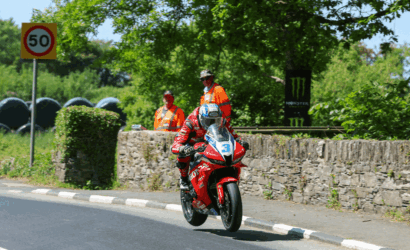 Motorcycle racer in red gear riding a red sport bike past a stone wall, number 3, with two orange-vested marshals watching.