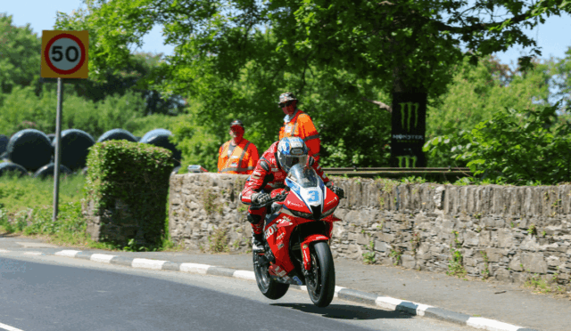 Motorcycle racer in red gear riding a red sport bike past a stone wall, number 3, with two orange-vested marshals watching.