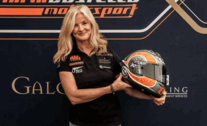 Woman in a black polo shirt holding a racing helmet, posing in front of a motorsport backdrop with logos.
