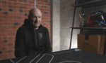 Older man in a black hoodie sits at a table in a garage, speaking to someone off-camera.