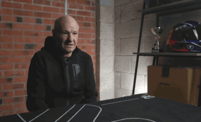 Older man in a black hoodie sits at a table in a garage, speaking to someone off-camera.