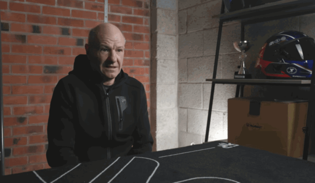 Older man in a black hoodie sits at a table in a garage, speaking to someone off-camera.