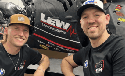 Two smiling men in black T-shirts sit beside a black race car with LEW Electrical Distributors branding in the background.