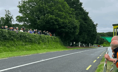 Spectators line a roadside barrier watching a race along a rural road with trees and hedges on the left.