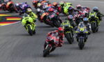 Group of motorcycle racers leaning into a sharp turn on a race track, lead rider in a bright red suit at the front of the pack.