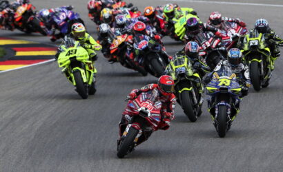 Group of motorcycle racers leaning into a sharp turn on a race track, lead rider in a bright red suit at the front of the pack.