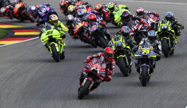 Group of motorcycle racers leaning into a sharp turn on a race track, lead rider in a bright red suit at the front of the pack.