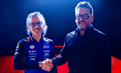 Two men shake hands on a blue-lit stage; one wears a branded race-team polo with sponsor logos and glasses, smiling at the camera.