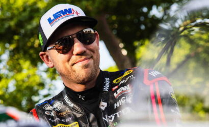 Smiling male racer wearing a cap with sponsor logo and sunglasses, in a black racing suit outdoor among trees.