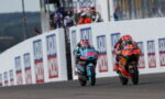 Two motorcycle racers sprint side by side on a race track, leaning into the turn past sponsor boards.